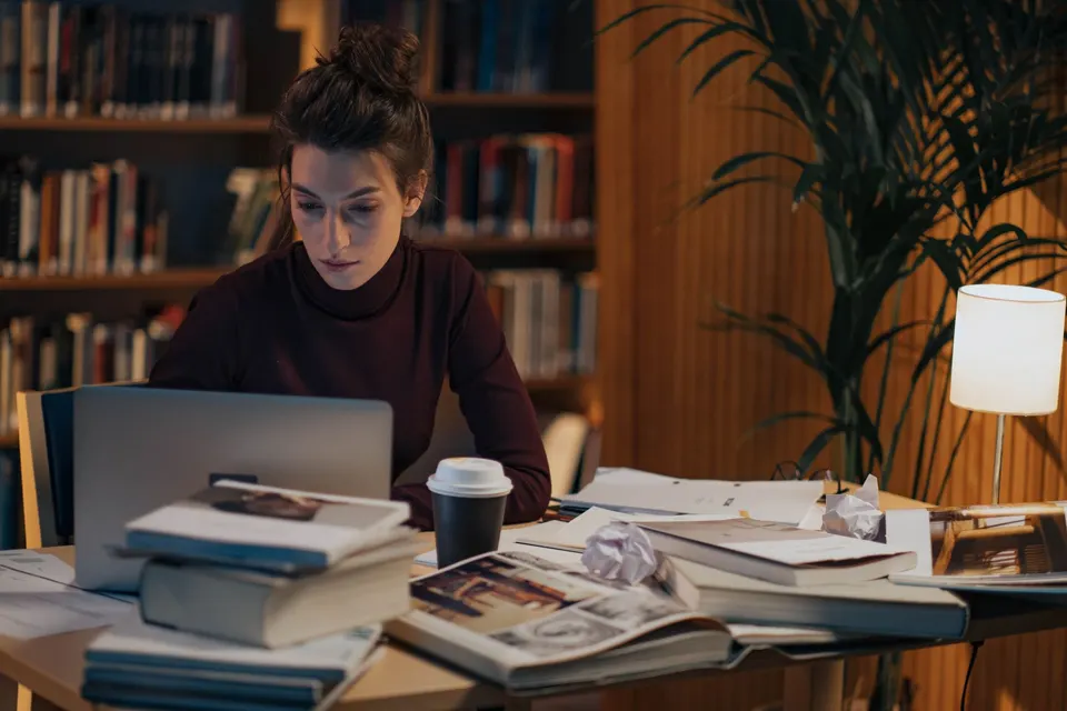 A student working late at a library desk covered in books and papers, with a laptop and coffee.