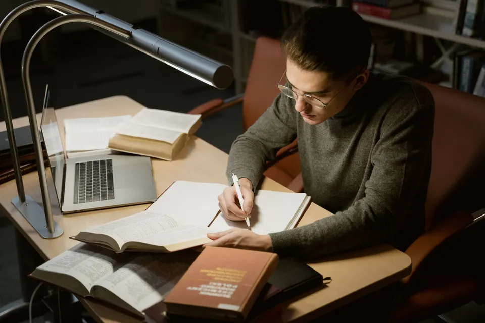 A student taking notes at a library desk under a lamp, with open books and a laptop.