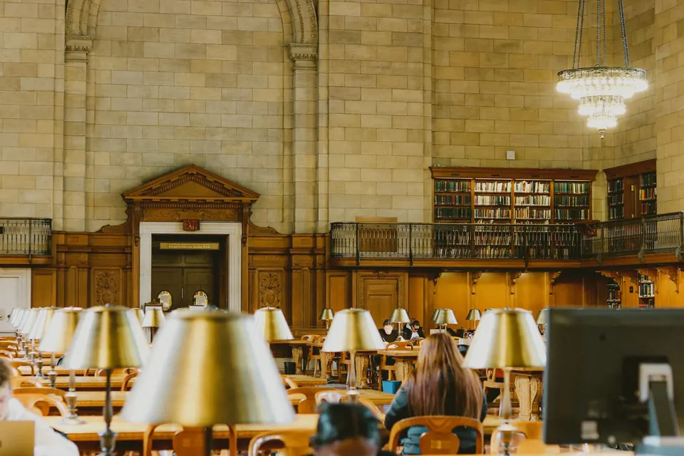 Students studying in a grand library reading room under chandeliers and brass lamps.