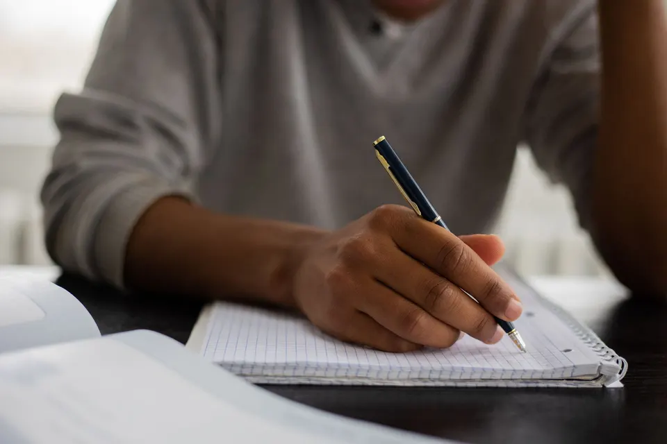A student writing notes in a workbook beside an open textbook.